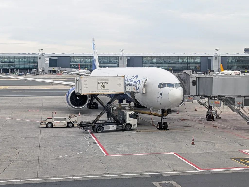 An IndiGo aeroplane is parked at an airport gate as a catering lorry services it and a jet bridge connects to the aircraft for boarding or disembarking.