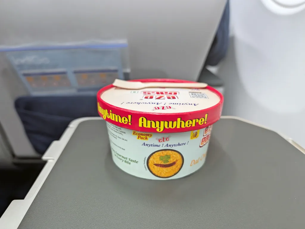 A cup of instant noodles with a wooden fork rests on an aeroplane tray table, with aeroplane seats and pocket materials visible in the background.