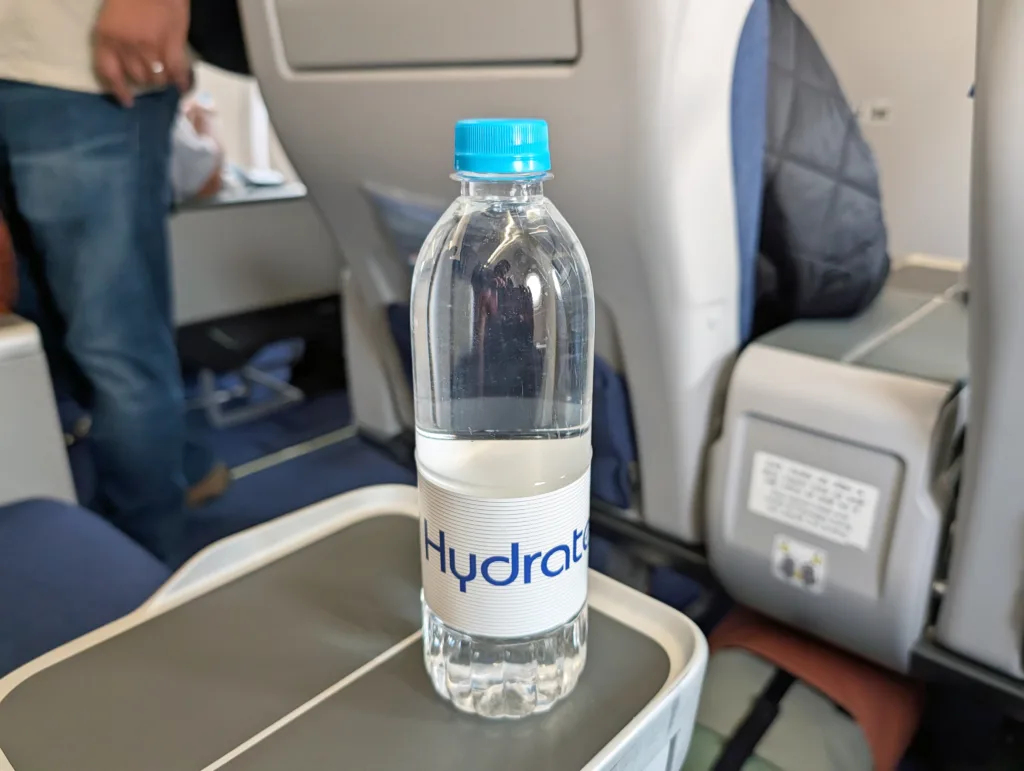 A clear plastic water bottle labelled "Hydrate" with a blue cap sits on a tray table in an aeroplane cabin.