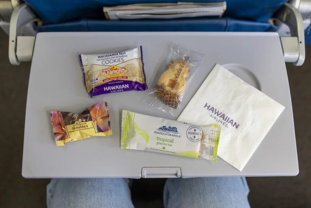 An aeroplane tray table with packaged snacks crafted by Hawaiian Airlines chef Sheldon Simeon, a serviette labelled "Hawaiian Airlines," and a passenger's legs partially visible below.