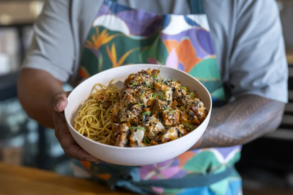 A person in a colourful apron holds a bowl of noodles topped with seasoned chicken pieces and garnished with herbs and sesame seeds.