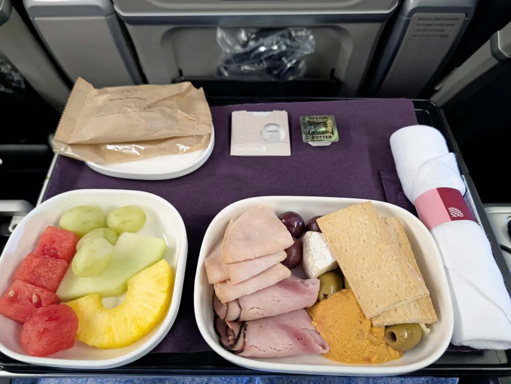 A tray of aeroplane food with sliced fruit, cold meats, cheese, biscuits, olives, a packet of butter, packaged biscuits, and a wrapped serviette on a seatback tray table.
