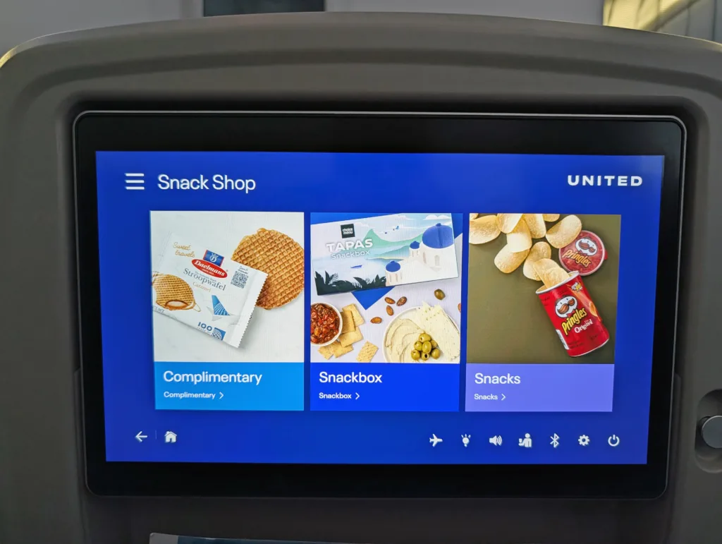 An in-flight entertainment screen displays three snack options: complimentary stroopwafel, a tapas snackbox, and Pringles crisps, with the United Airlines logo in the top right.