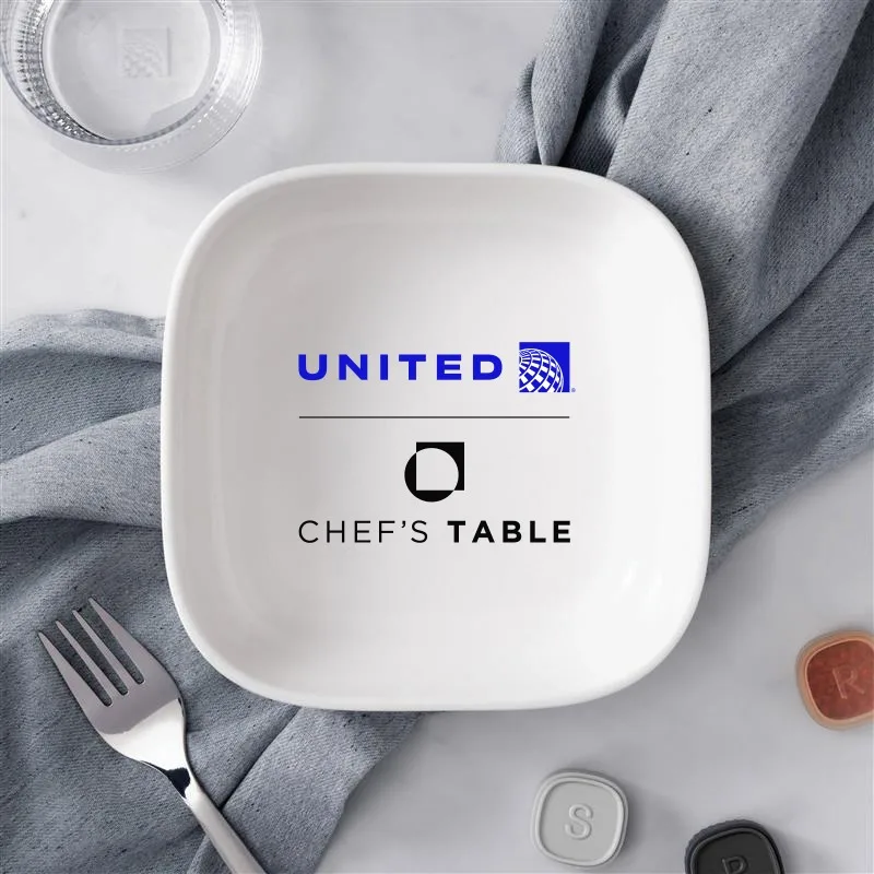 United Airlines launches Chef's Table collaboration August 2026 2 A white square dish with the United Airlines and Chefโs Table logos printed inside, placed on a grey cloth with a fork nearbyโnot to be confused with JetBlue or Cown Shy branding.